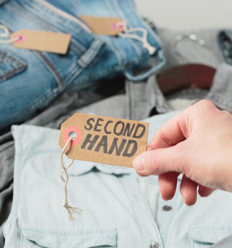 Clothes from females wardrobe on shelf of second hand store. Womans hand holding craft paper tag with inscription second hand. Circular fashion, eco friendly shopping, thrift stores concept