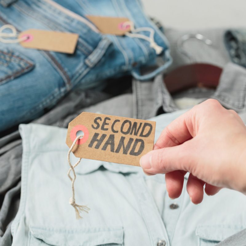 Clothes from females wardrobe on shelf of second hand store. Womans hand holding craft paper tag with inscription second hand. Circular fashion, eco friendly shopping, thrift stores concept
