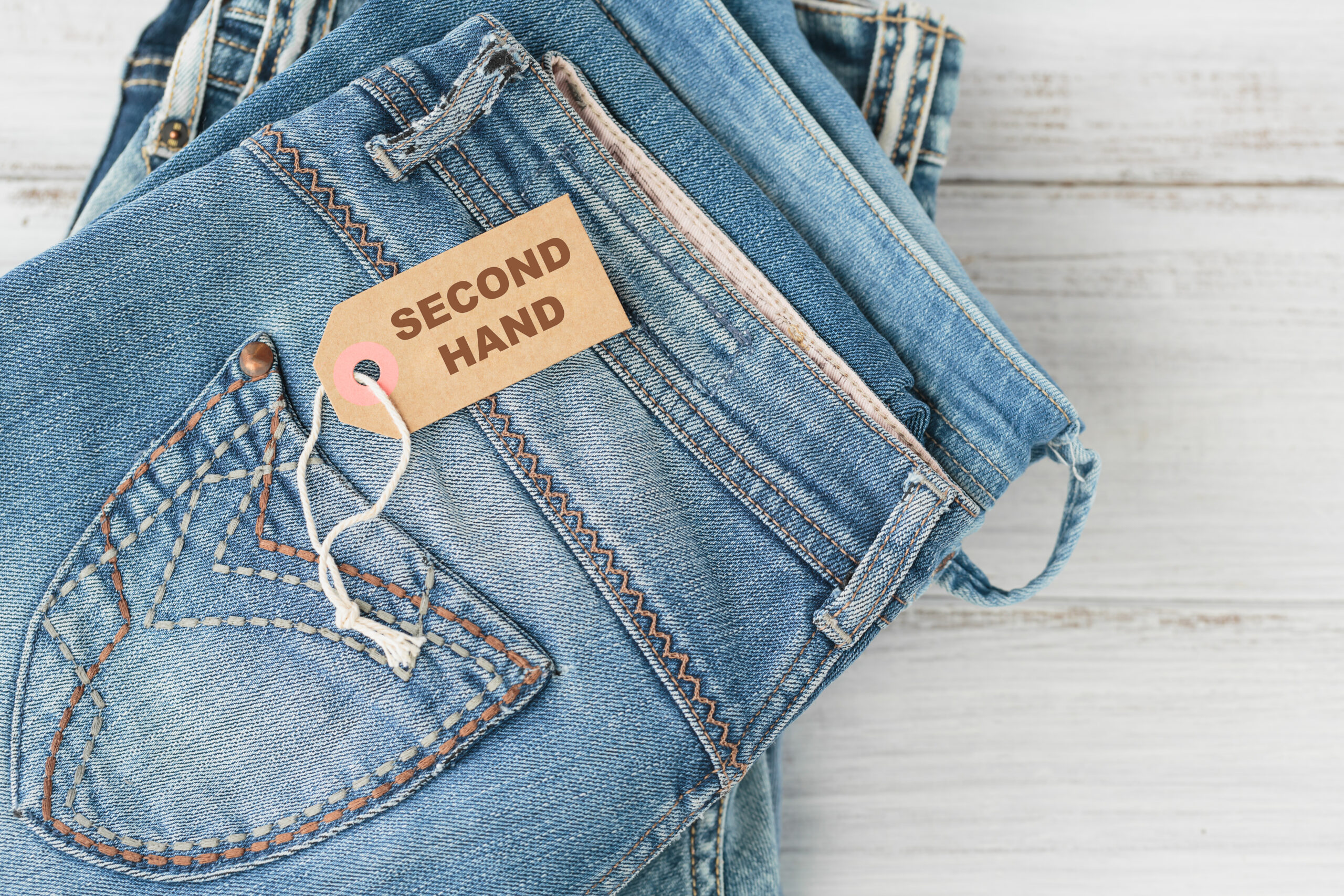 Stack of blue jeans and tag with inscription second hand on white background. Second hand clothing shop. Circular fashion, eco friendly sustainable shopping, thrift stores concept