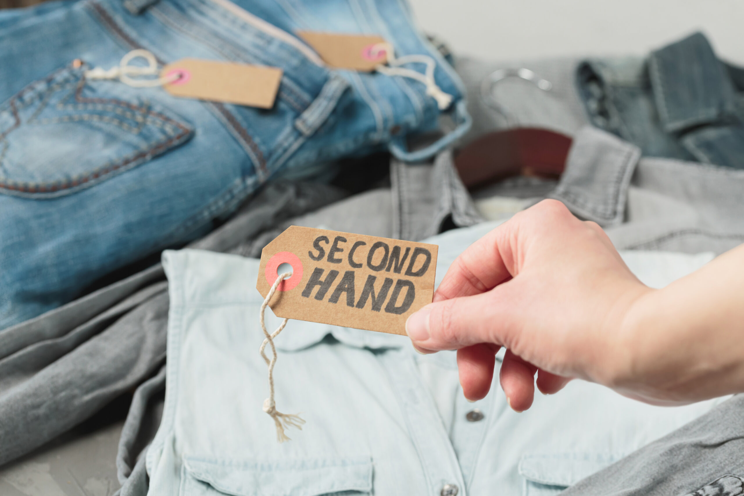 Clothes from females wardrobe on shelf of second hand store. Womans hand holding craft paper tag with inscription second hand. Circular fashion, eco friendly shopping, thrift stores concept